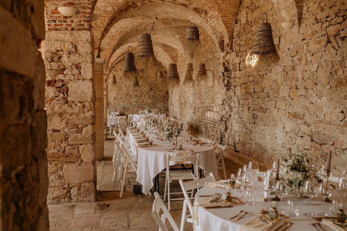 Long tables with ivory florals and rattan pendants in vaulted stone cellar with rounded brick arches