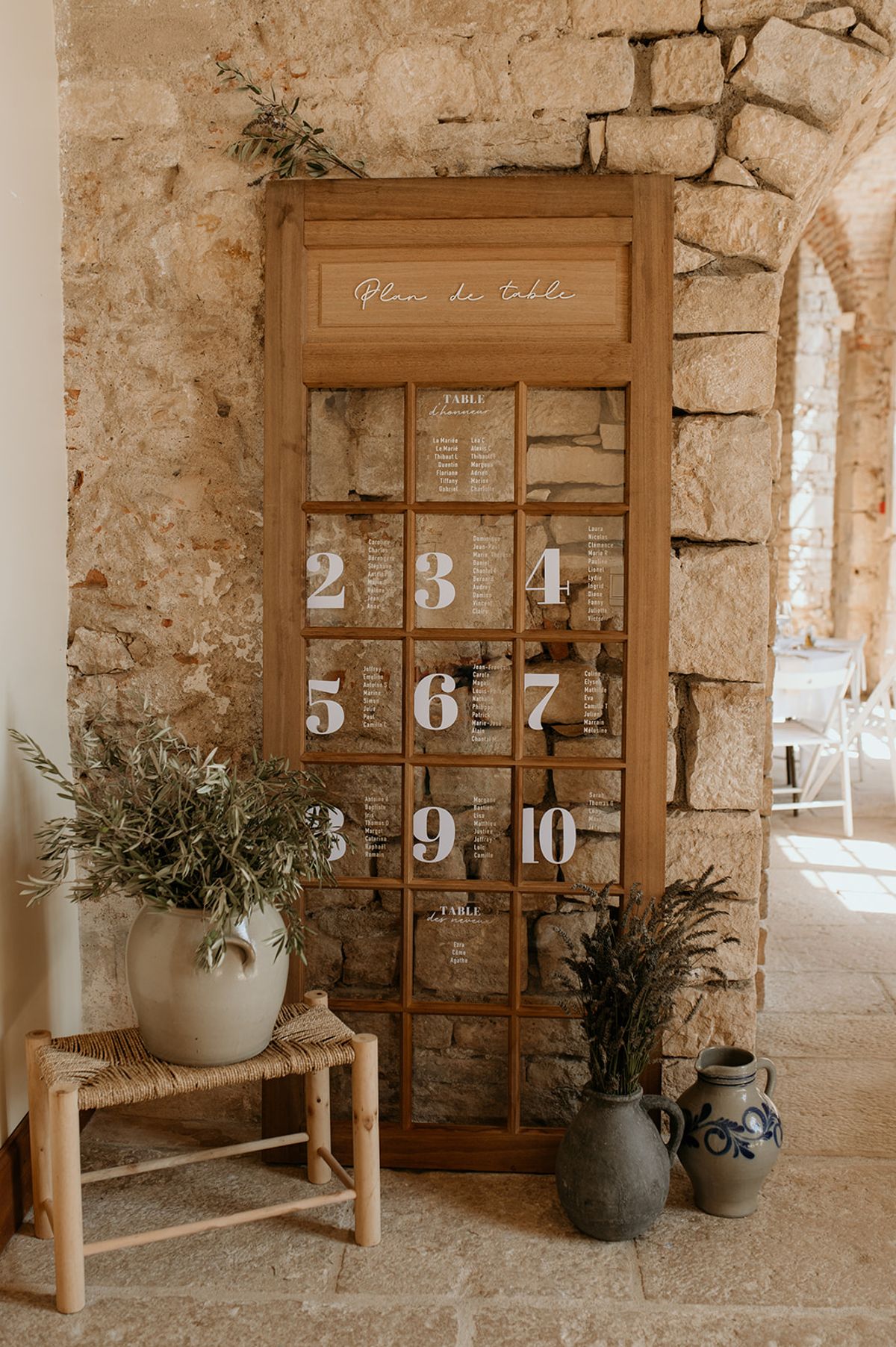 Rustic seating chart on repurposed wooden window frame with olive branches and stoneware jugs under stone arch