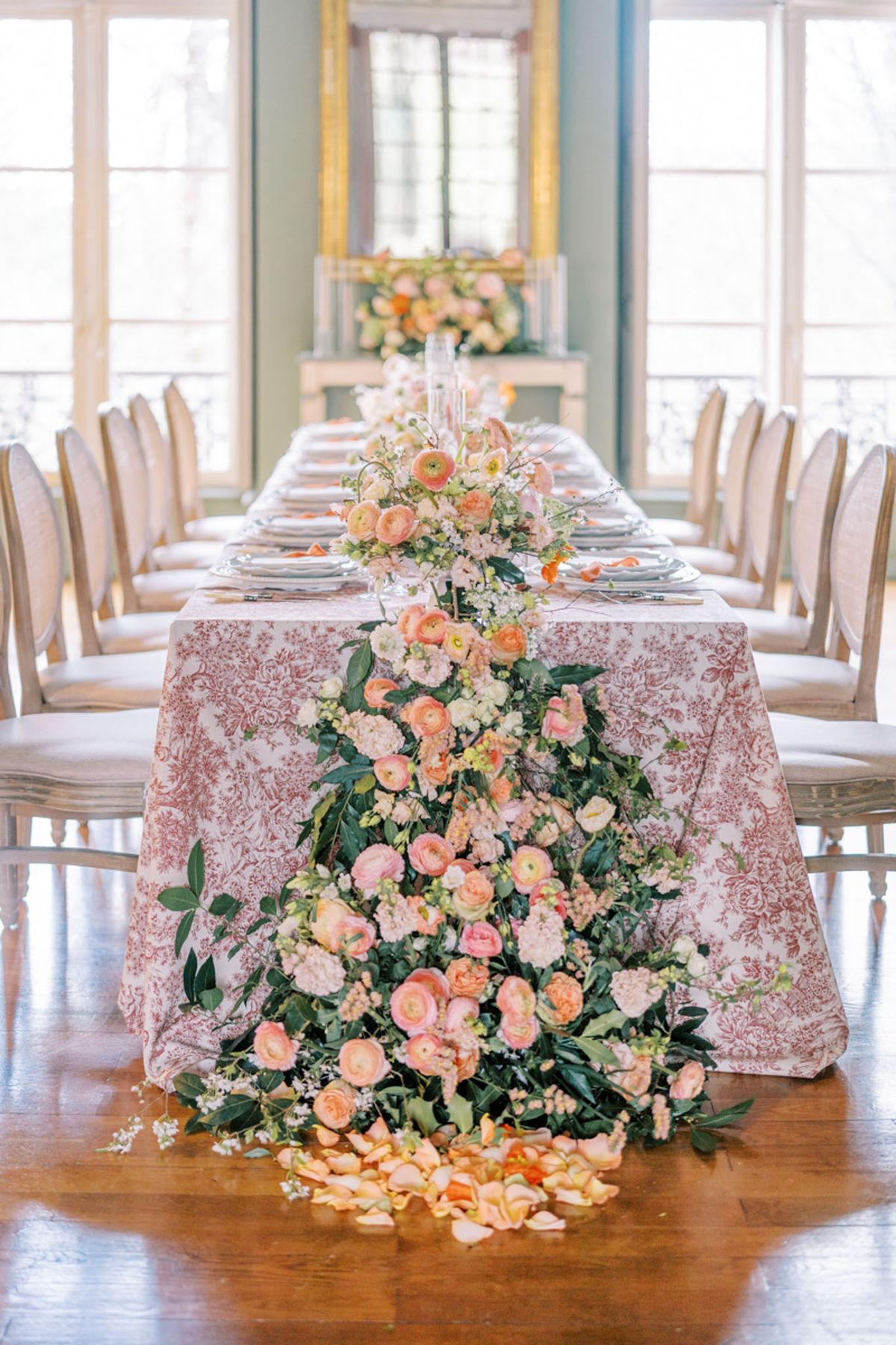 A reception tablescape detail shot taken indoors in a classical French-style room with pale sage-painted walls, tall windows, and a gilt-framed mirror. The long rectangular dining table is covered in a dusty rose toile de Jouy printed tablecloth and set with silver charger plates, flatware, and place settings for approximately 14 guests, with French linen-upholstered wood chairs on either side. The central floral arrangement is a dramatic cascading installation of peach and coral ranunculus, blush stock flowers, garden roses, white wisteria-like blooms, and mixed greenery that runs along the top of the table and spills down the end, pooling onto the hardwood floor with scattered orange and peach rose petals. A tall glass candlestick holder is visible at the center of the table, and a secondary floral arrangement in peach and cream tones sits on a console near the mirror in the background. The overall design palette is coral, peach, blush, and green with a classic French aesthetic.