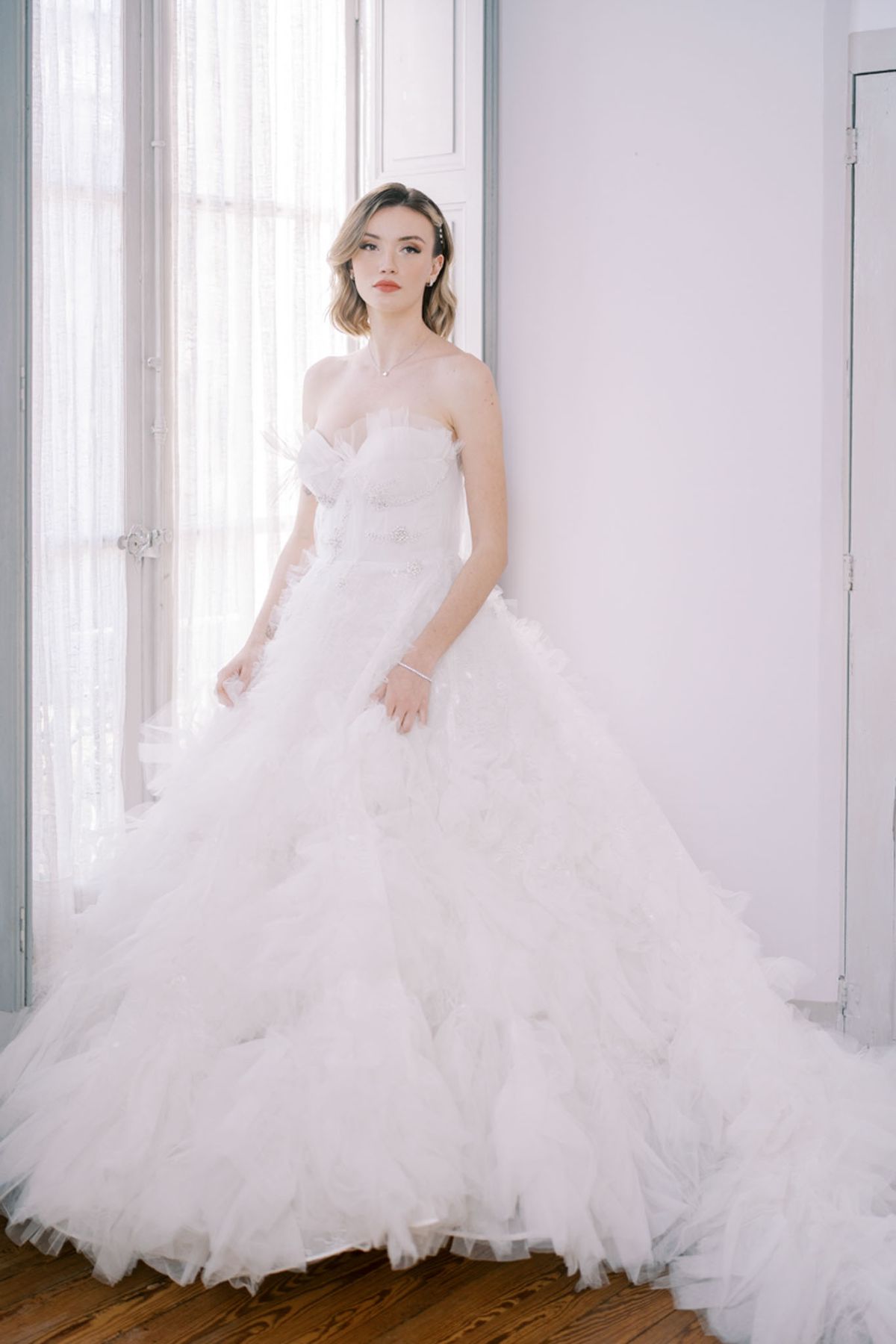 A bridal portrait of a single woman photographed indoors in a bright, white-walled room with tall French windows dressed in sheer white curtains. She wears a strapless white ball gown with a fitted, beaded bodice featuring floral appliqué detailing and a voluminous, multi-layered ruffled tulle skirt with a full train that spreads across the hardwood floor. Her hair is styled in a soft, short wave with pearl or crystal hair pins, and she wears a delicate pendant necklace, small earrings, and a bracelet. Her makeup includes a bold coral-red lip. The shot is a full-length portrait with soft, diffused natural light coming from the window behind her, creating a bright, airy, high-key aesthetic consistent with a classic or editorial bridal style.