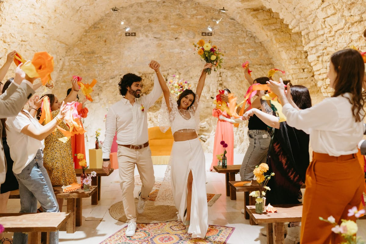 Couple recessioning through stone cave venue with colourful crepe paper waves coral bridesmaids and bohemian rugs