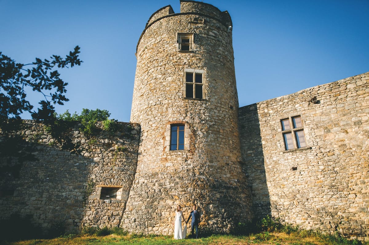 Bride and groom hand-in-hand dwarfed by medieval stone tower in golden-hour light