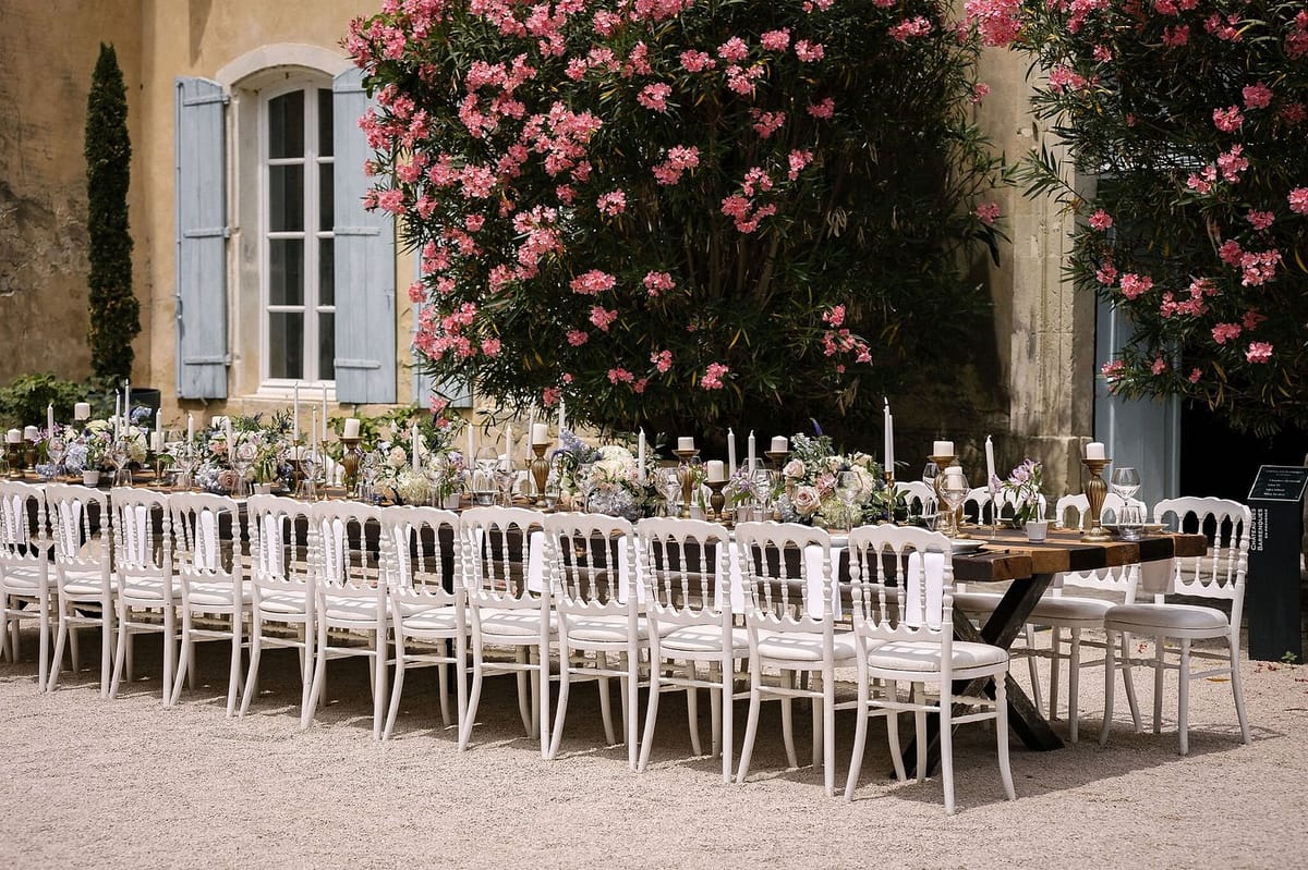 An outdoor wedding reception table setup photographed in wide shot against the façade of a Provençal-style mas or château with pale yellow rendered walls and blue-grey shuttered windows. A single long feasting table with a dark wood top and cross-leg base is lined on both sides with white Napoleon-style chairs with upholstered cream seats, seating approximately 20–24 guests. The table is dressed with a runner of low floral arrangements featuring blush roses, dusty blue hydrangeas, ivory blooms, and greenery, interspersed with brass candelabras and single white taper candles in gold holders. Glassware and place settings are visible along the length of the table. The overall decor palette combines warm gold, white, blush, and muted blue tones, giving a classic French countryside aesthetic. Potential venue feature image.