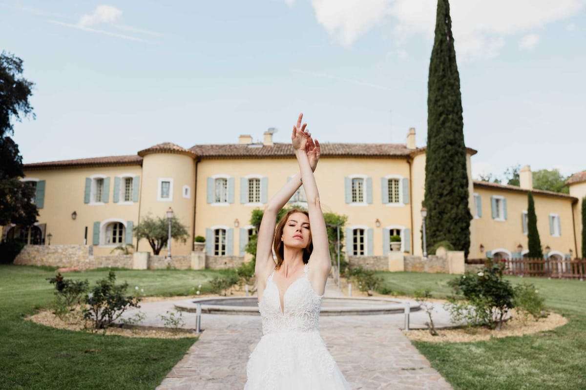 A bride poses outdoors in the formal garden of a Provençal-style manor house, standing in front of a circular stone fountain with her arms raised overhead in a balletic pose, eyes closed. She wears a white lace A-line wedding dress with a deep V-neckline, thin spaghetti straps, and a textured layered skirt. The venue behind her is a large yellow-rendered two-storey manor with pale teal-blue shutters, a terracotta tiled roof, and rounded corner towers, flanked by tall cypress trees. This is a full-length portrait shot with the bride as the sharp focal point and the manor softly blurred in the background. Potential venue feature image.