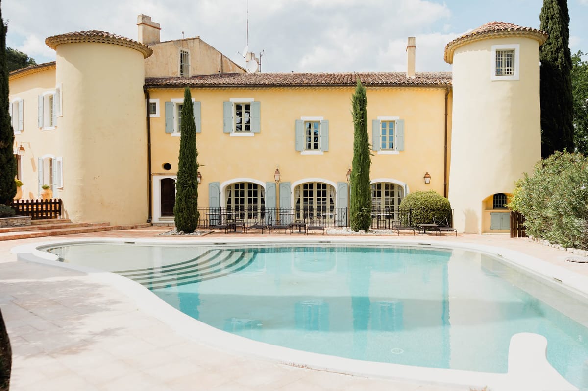 Wide exterior shot of a Provençal-style mas or bastide, featuring warm yellow ochre rendered walls, pale blue-grey shutters, arched French doors, terracotta roof tiles, and two rounded corner towers. The façade is flanked by tall columnar cypress trees and copper wall lanterns. A large kidney-shaped swimming pool with pale stone coping occupies the foreground, with wrought-iron chairs and small tables arranged on the terrace along the ground-floor arched openings. No people are visible in the image. Potential venue feature image.