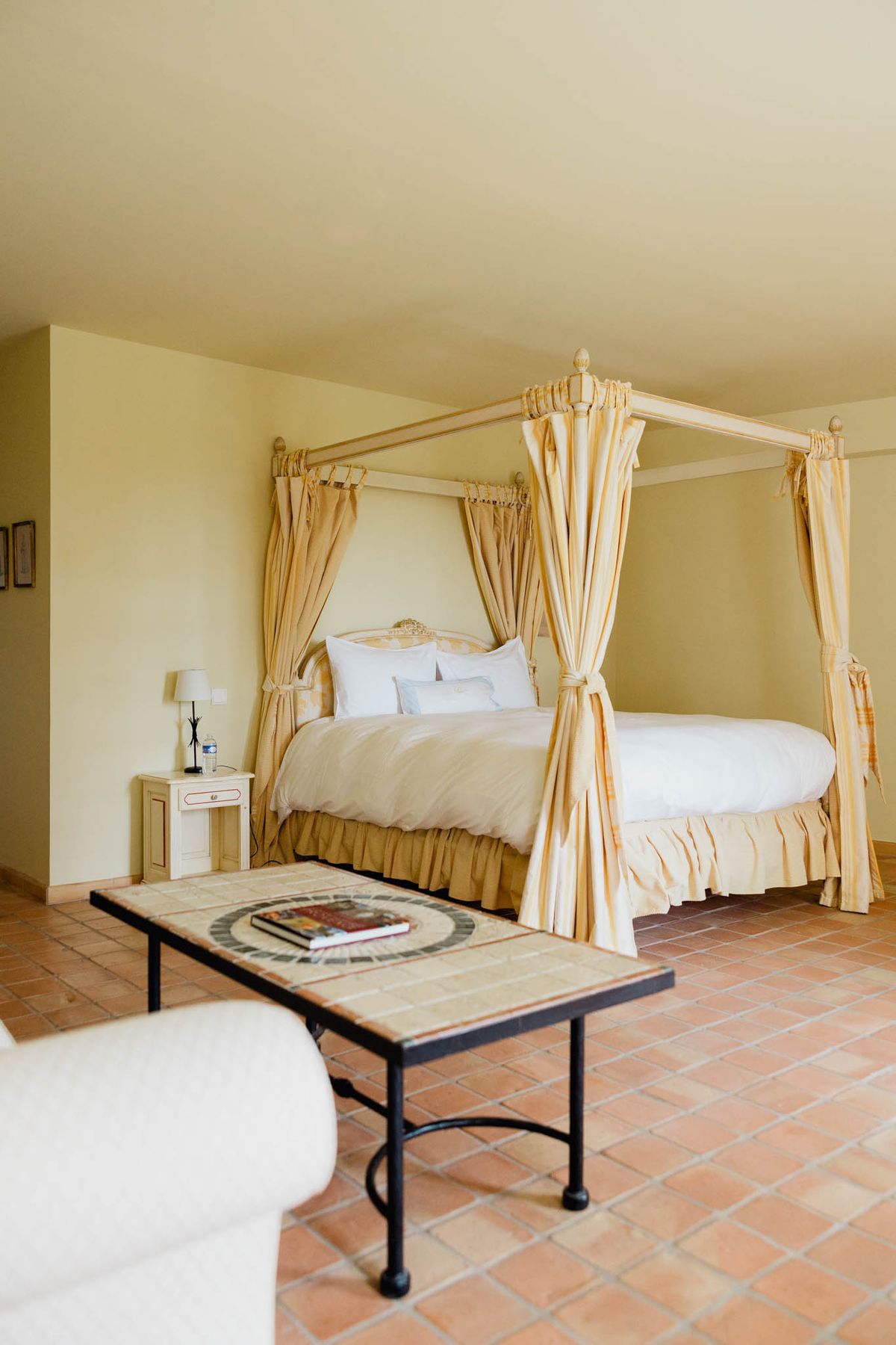 Wide interior shot of a hotel or chateau bedroom, likely used as a bridal suite or getting-ready space for a wedding. The room features a four-poster canopy bed with a carved wooden frame and gold-and-cream striped drapes tied back at each post, dressed with white linens and multiple white pillows. A cream-painted nightstand with a small lamp sits to the left of the bed. The flooring is terracotta tile, and a wrought-iron coffee table with a mosaic tile top and a couple of books sits in the foreground, alongside the edge of a cream upholstered armchair. The walls are painted a warm pale yellow, giving the room a classic Provençal style. No people are present. Potential venue feature image.