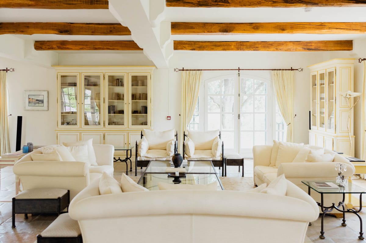 French country salon with exposed beams cream sofas glazed armoire cabinets wrought-iron tables and arched French doors