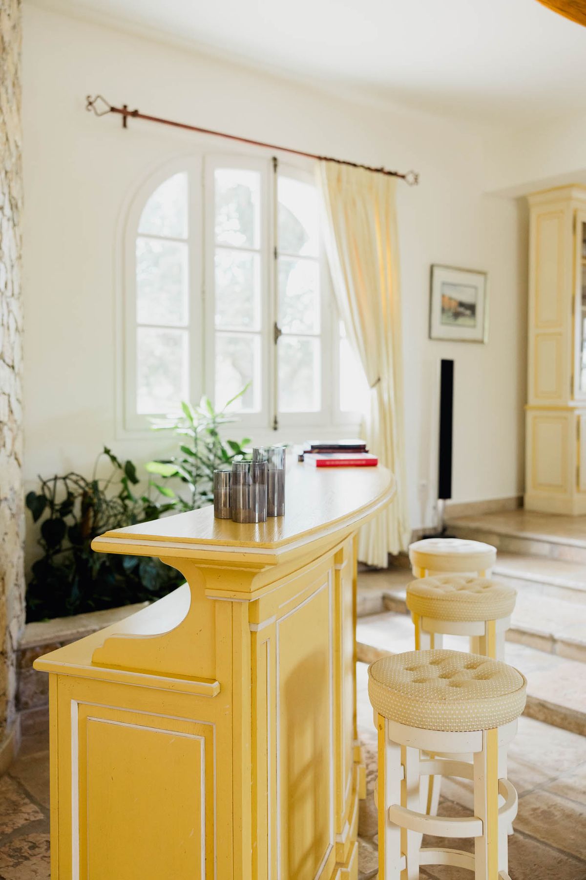 Provencal interior bar with mustard yellow counter cream tufted stools arched window and striped curtains