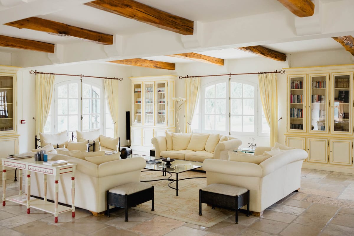 Spacious salon with exposed wood beams, cream sofas, wrought iron coffee table, and built-in bookcases in a French country...