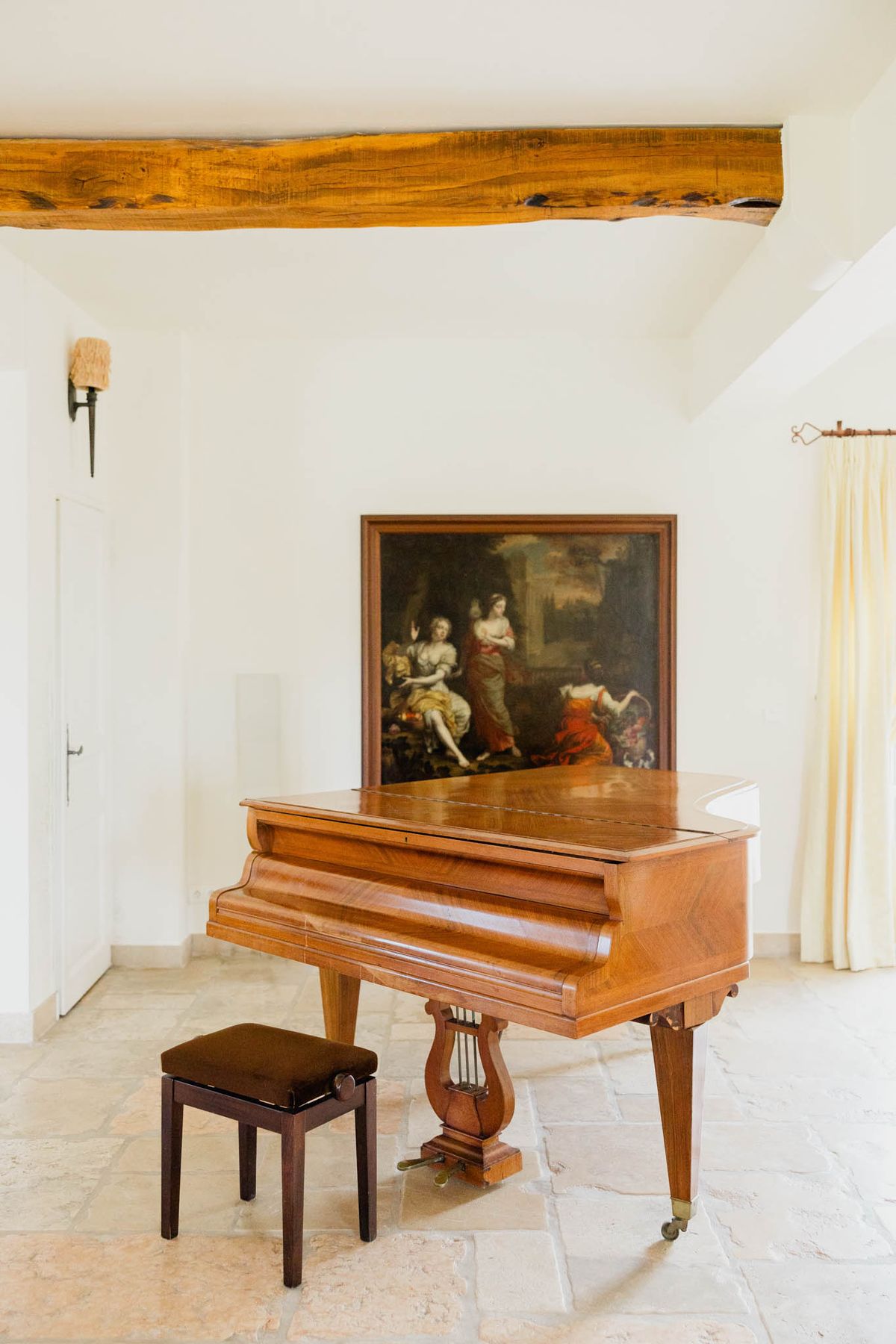 An interior detail shot of a venue room featuring a warm honey-toned walnut baby grand piano with a lyre-shaped pedal support and brass caster feet, accompanied by a dark wood upholstered bench. Behind the piano sits a large framed Old Master-style oil painting depicting several female figures in classical dress against a dark background. The room has white plastered walls, a rustic exposed wooden ceiling beam, stone tile flooring in a warm cream tone, cream linen curtains on the right, and a single wall-mounted sconce on the left. The overall decor style blends classic French country with antique furnishings. Wide shot, interior perspective. Potential venue feature image.