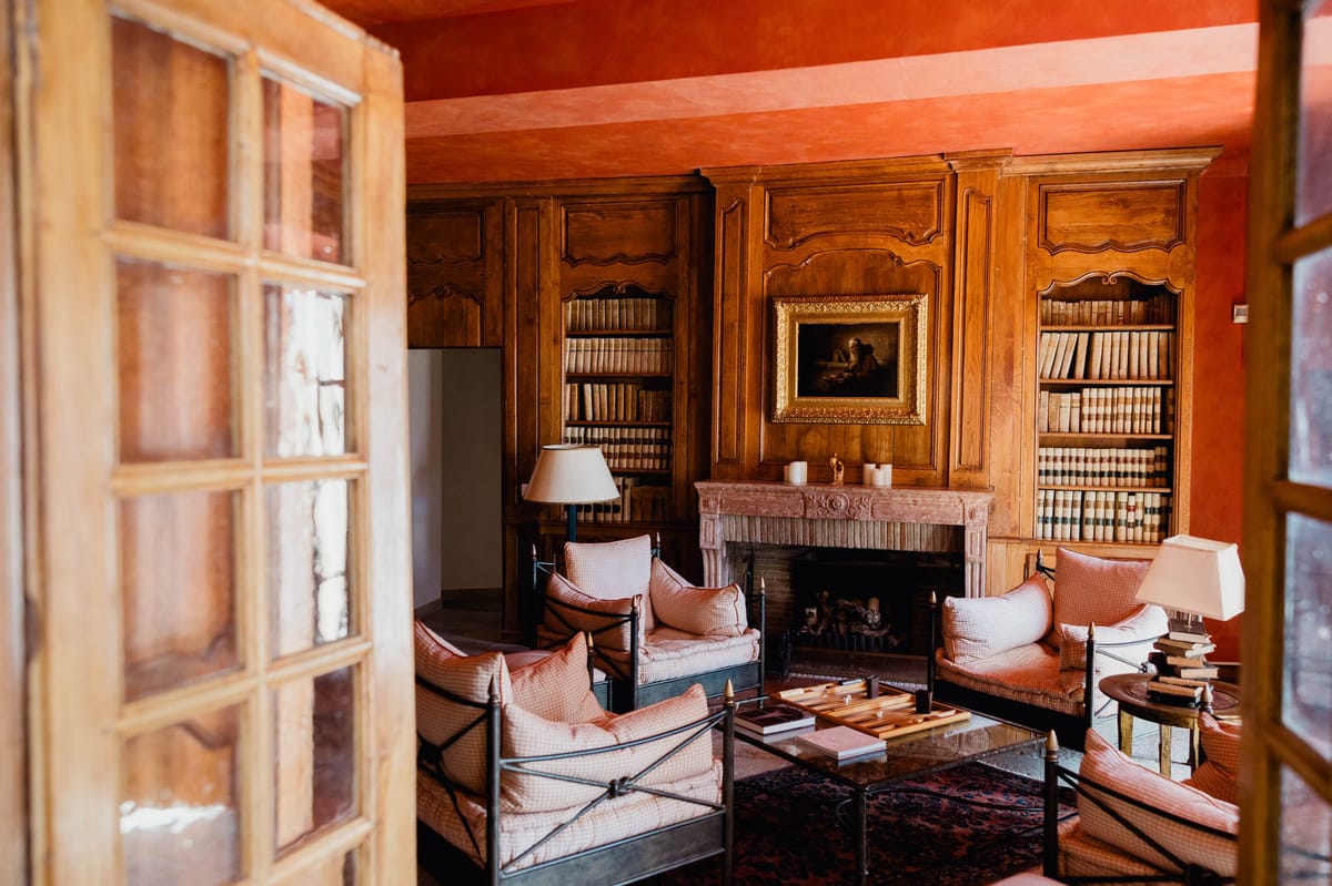 An interior room of a French château or manor house, photographed through open wooden French doors with glass panes. The room features deep terracotta-painted walls and ceiling, floor-to-ceiling carved walnut boiserie panels with integrated bookshelves lined with antique leather-bound volumes. A carved stone fireplace in blush-pink marble sits at the center of the room, topped with white pillar candles and a small figurine. Above the fireplace hangs an oil painting in a gilded gold frame depicting a figure at a desk. The seating area consists of several iron-framed armchairs and a sofa upholstered in dusty rose and cream checked fabric, arranged around a glass-topped coffee table with a backgammon set. A patterned burgundy and navy rug anchors the seating grouping, and two cream-shaded table lamps provide warm ambient lighting. The composition is a wide interior shot taken from the doorway threshold. Potential venue feature image.