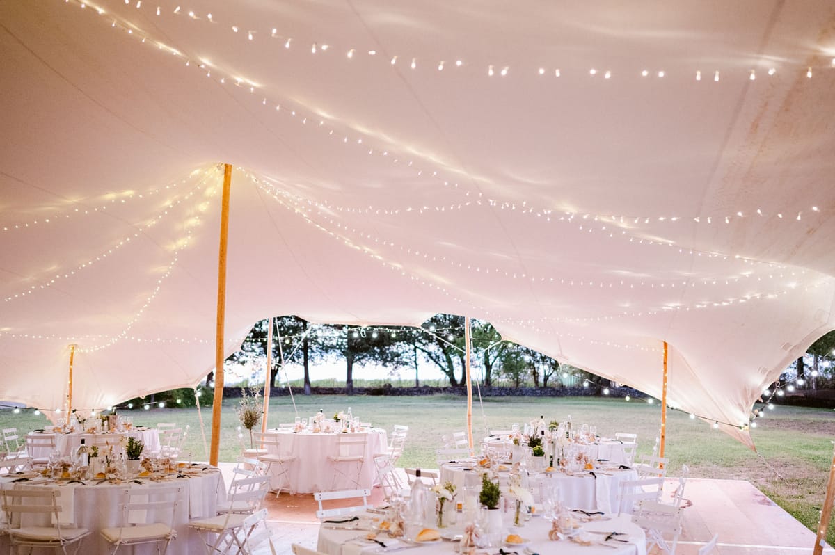 Stretch tent reception with fairy lights, round and long tables, and cream bud-vase florals at dusk