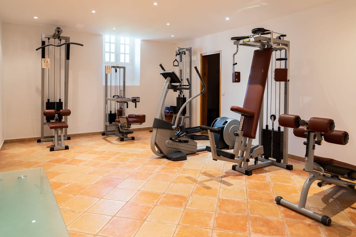 This image does not show a wedding photograph. It depicts an indoor fitness room or gym space featuring multiple pieces of exercise equipment including a cable lat pulldown machine, a seated rowing or leg press station, an elliptical trainer branded 'Excite', a multi-station cable weight machine with brown leather padding, and what appears to a decline bench. The room has terracotta-tiled flooring, white walls, recessed ceiling spotlights, and a single window providing natural light. No people, wedding-related subjects, or wedding decor are present in this image.