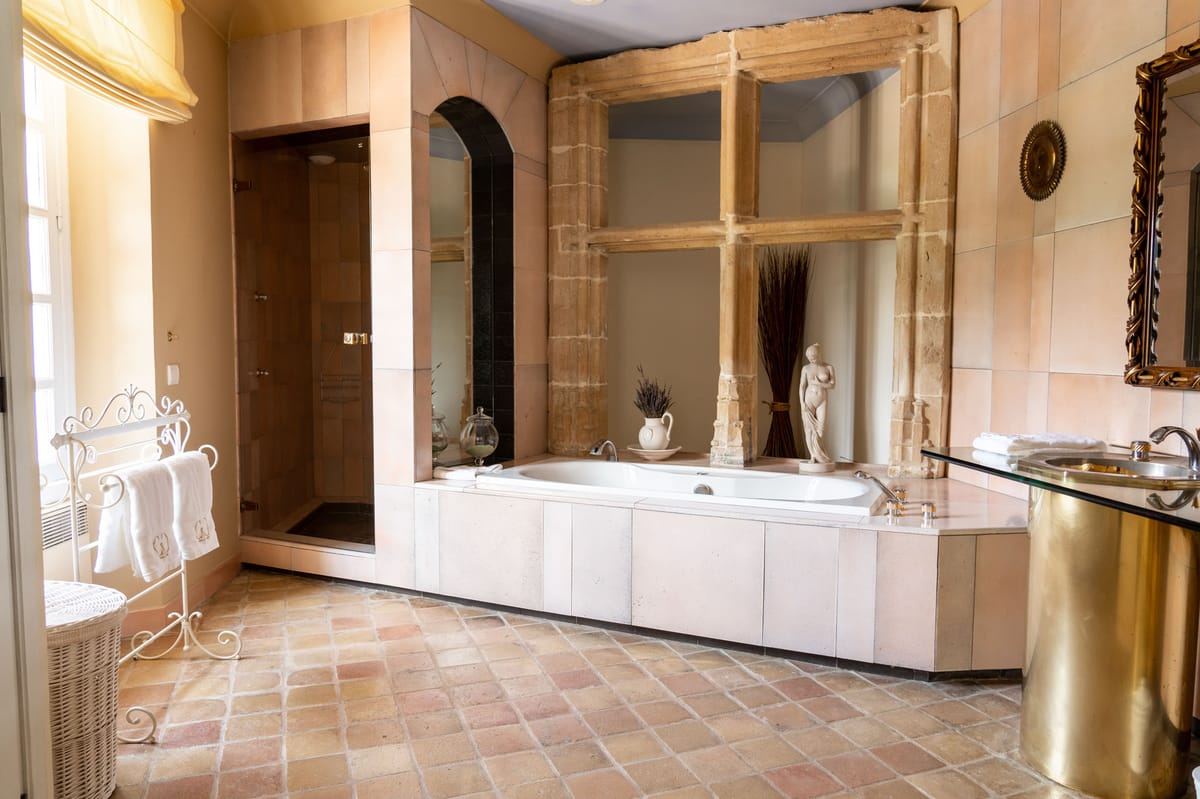 Chateau bathroom with Gothic stone columns framing a built-in bathtub, terracotta floor tiles, and brass vanity