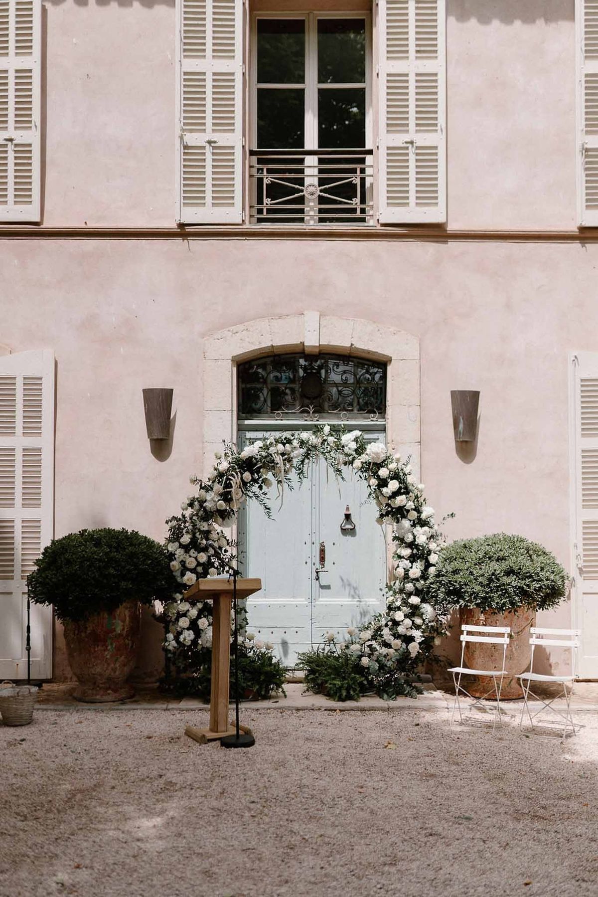 White floral ceremony arch framing blue doors on pink façade at Château Sainte-Roseline