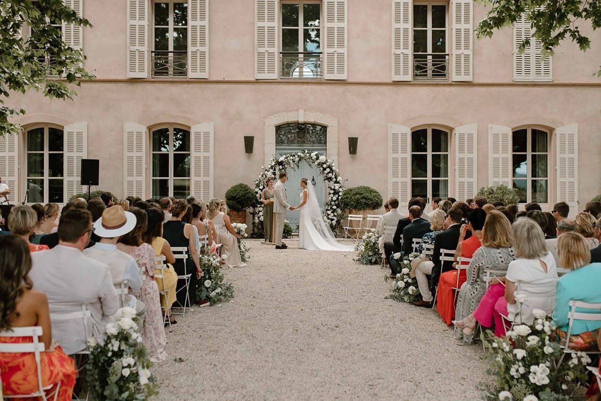 Couple at ceremony arch in courtyard with guests and pink façade at Château Sainte-Roseline