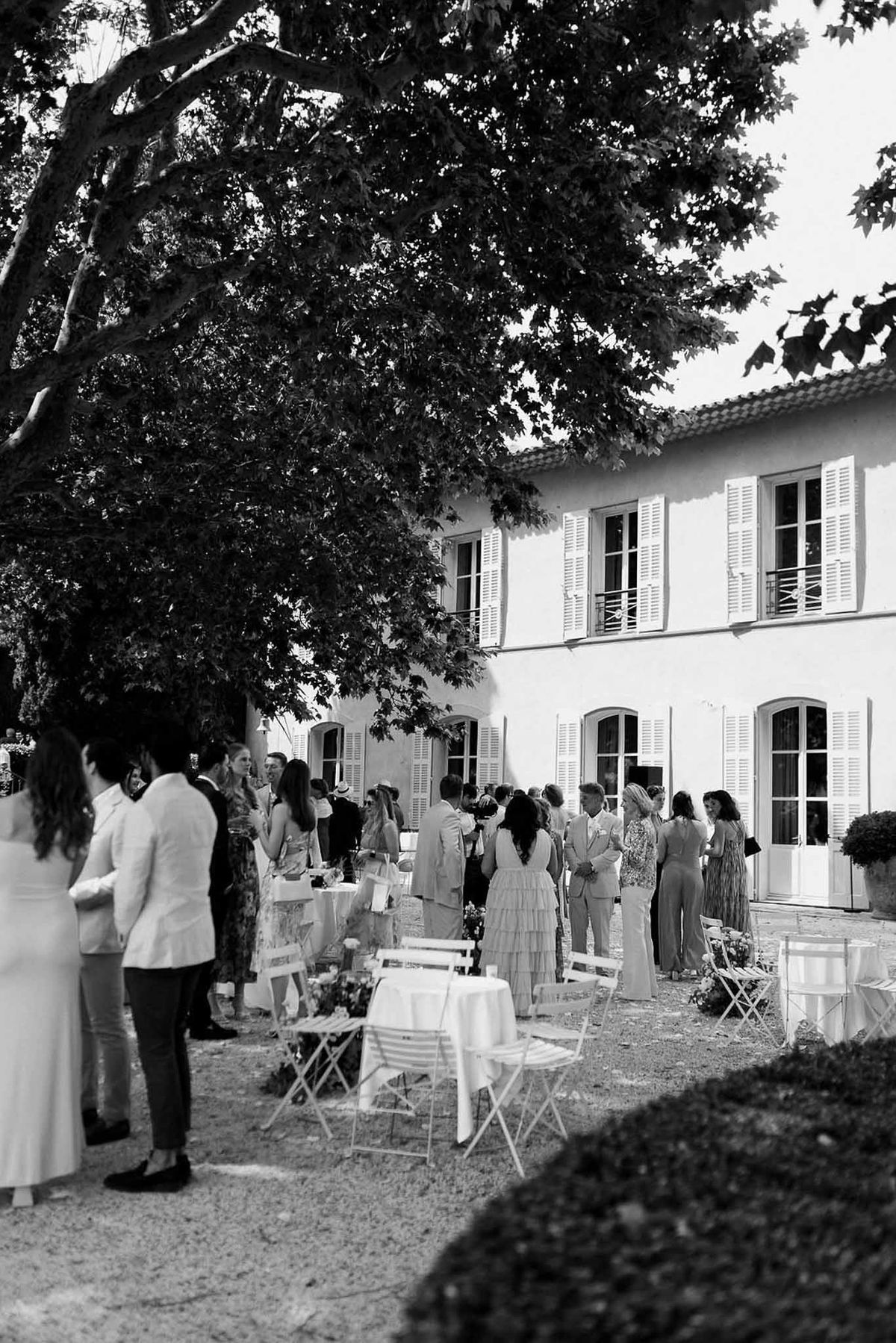 Guests at cocktail hour under plane tree beside château in black and white at Château Sainte-Roseline