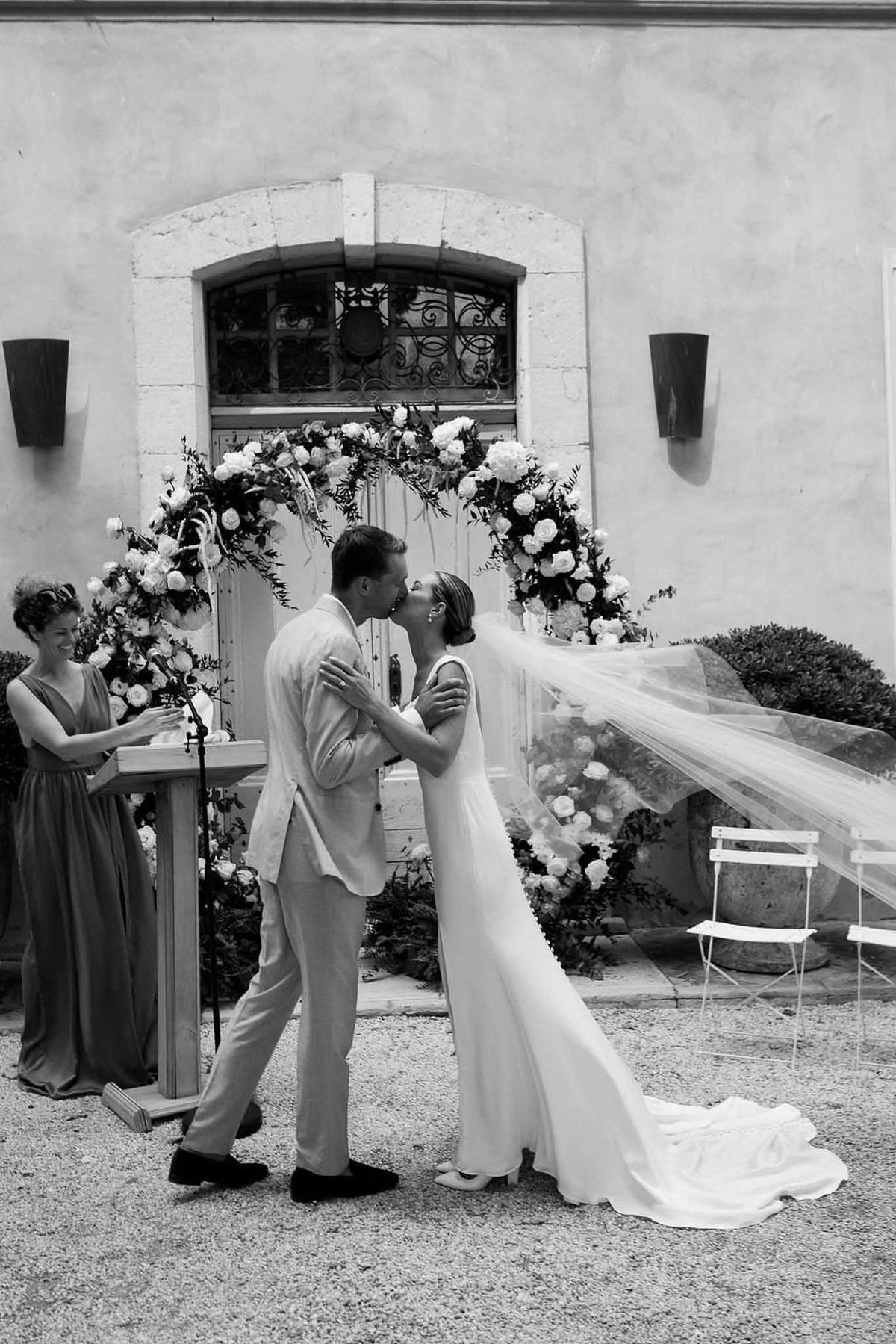 Couple's first kiss at floral arch with veil in breeze in black and white at Château Sainte-Roseline
