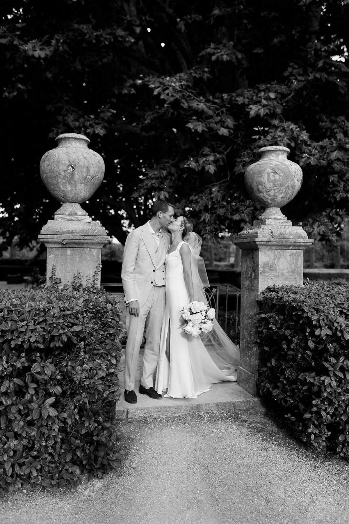 Couple kissing between stone urns on garden path in black and white at Château Sainte-Roseline