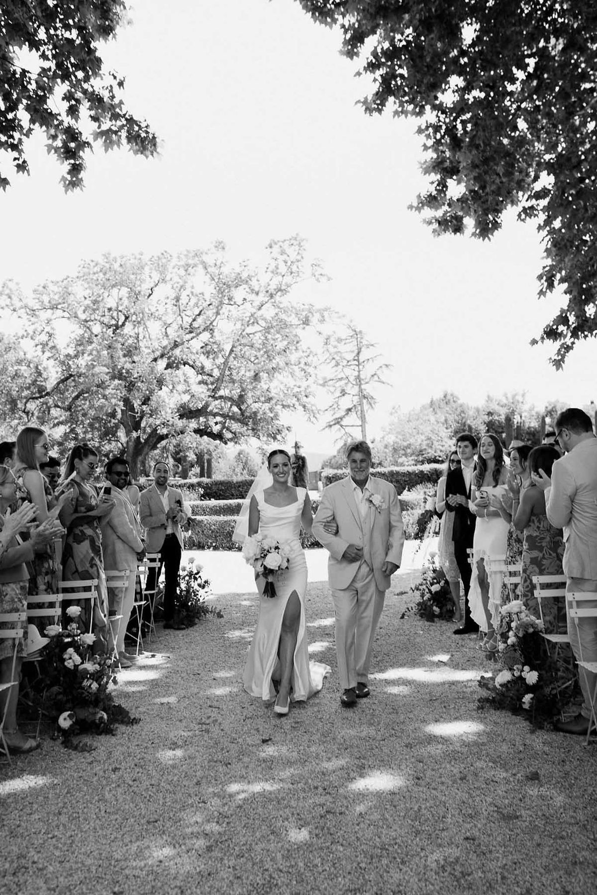 Bride and father walking down tree-lined aisle in black and white at Château Sainte-Roseline