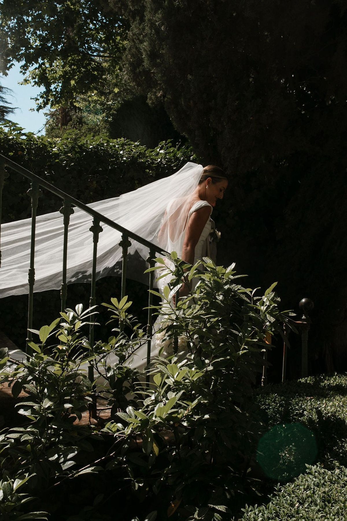 Bride descending wrought iron staircase with cathedral veil trailing through garden at Château Sainte-Roseline