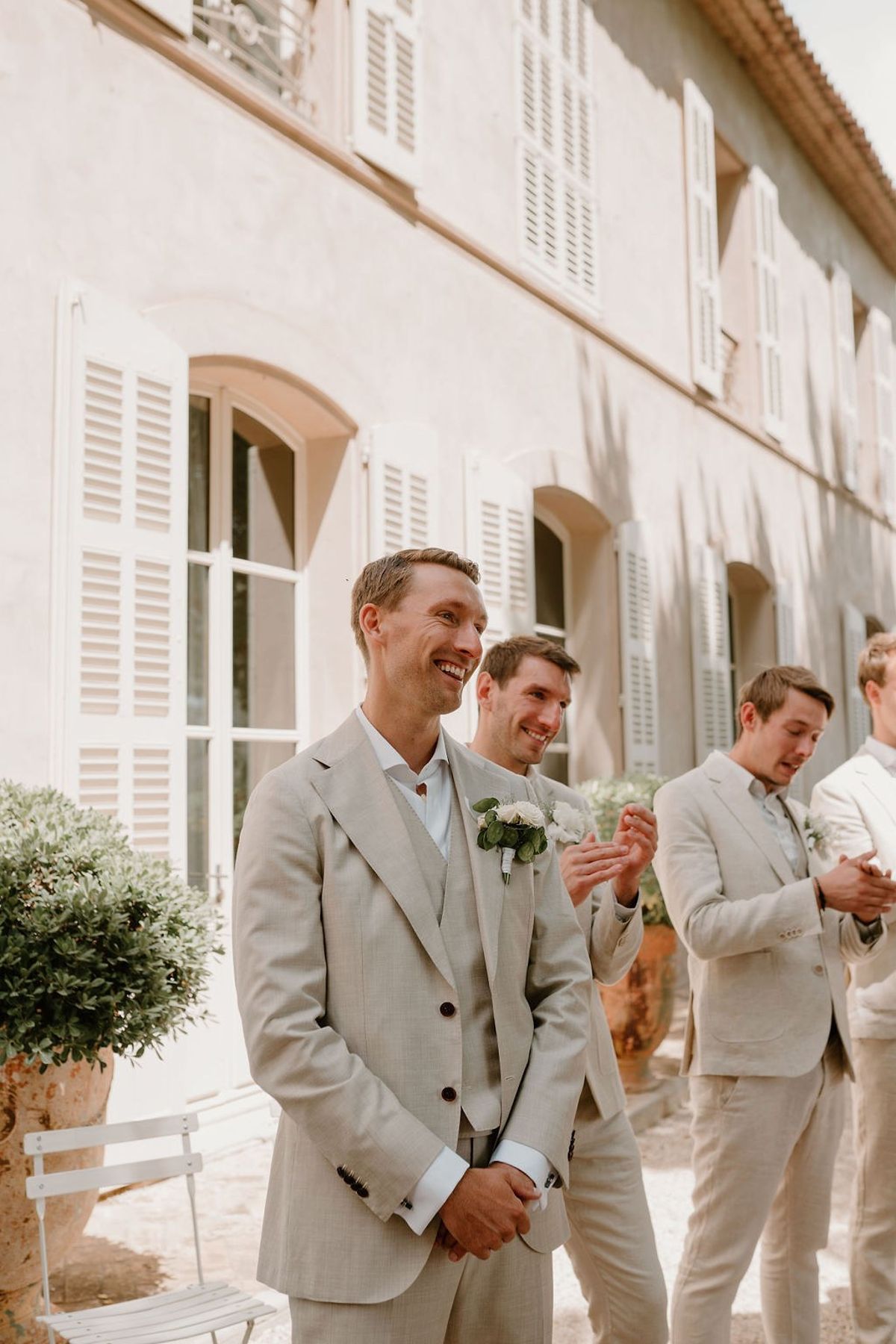 Groom and groomsmen smiling in linen suits before pink rendered facade and shutters at Château Sainte-Roseline