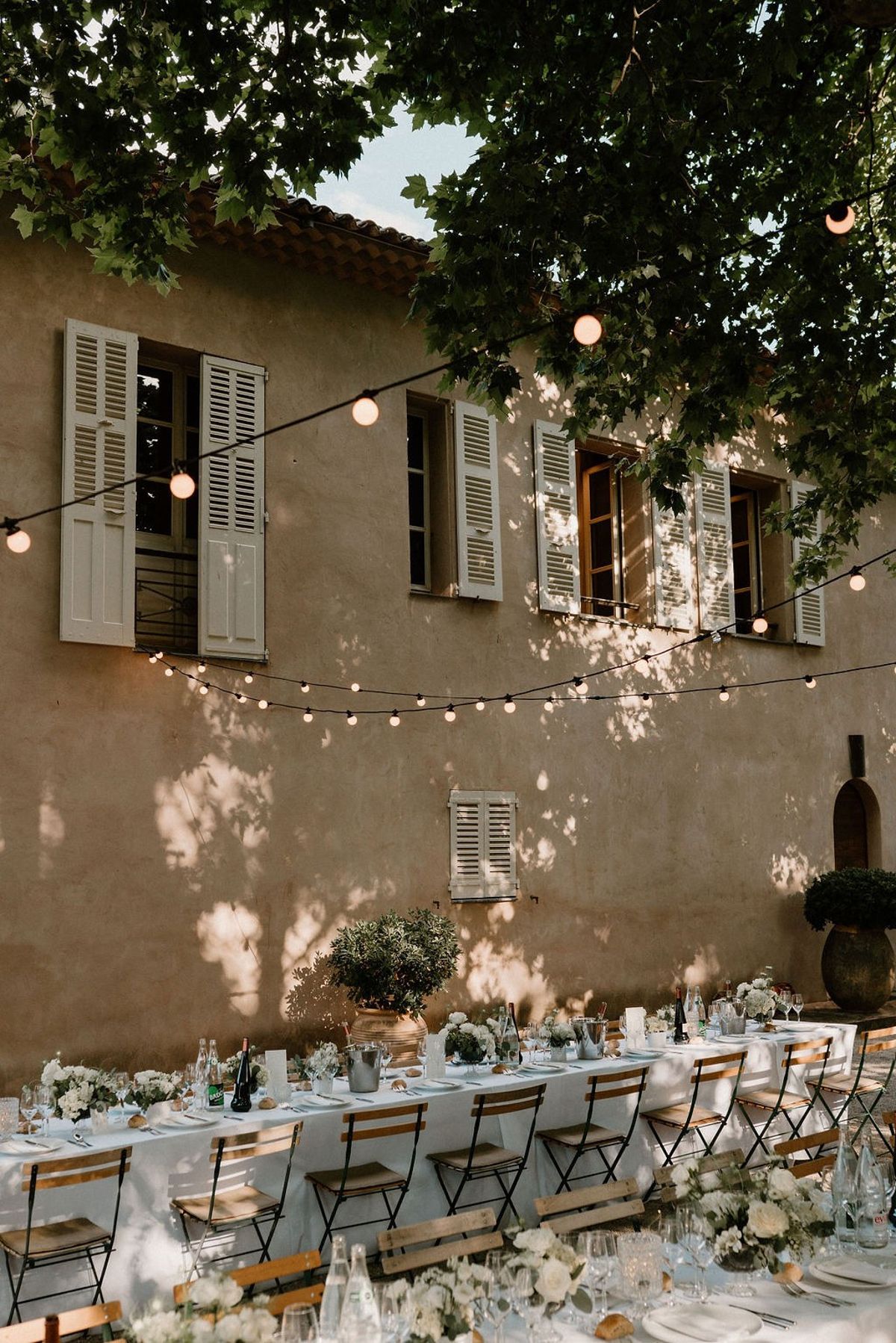 Long banquet table set with white florals and festoon lights against pink rendered chateau wall at Château Sainte-Roseline