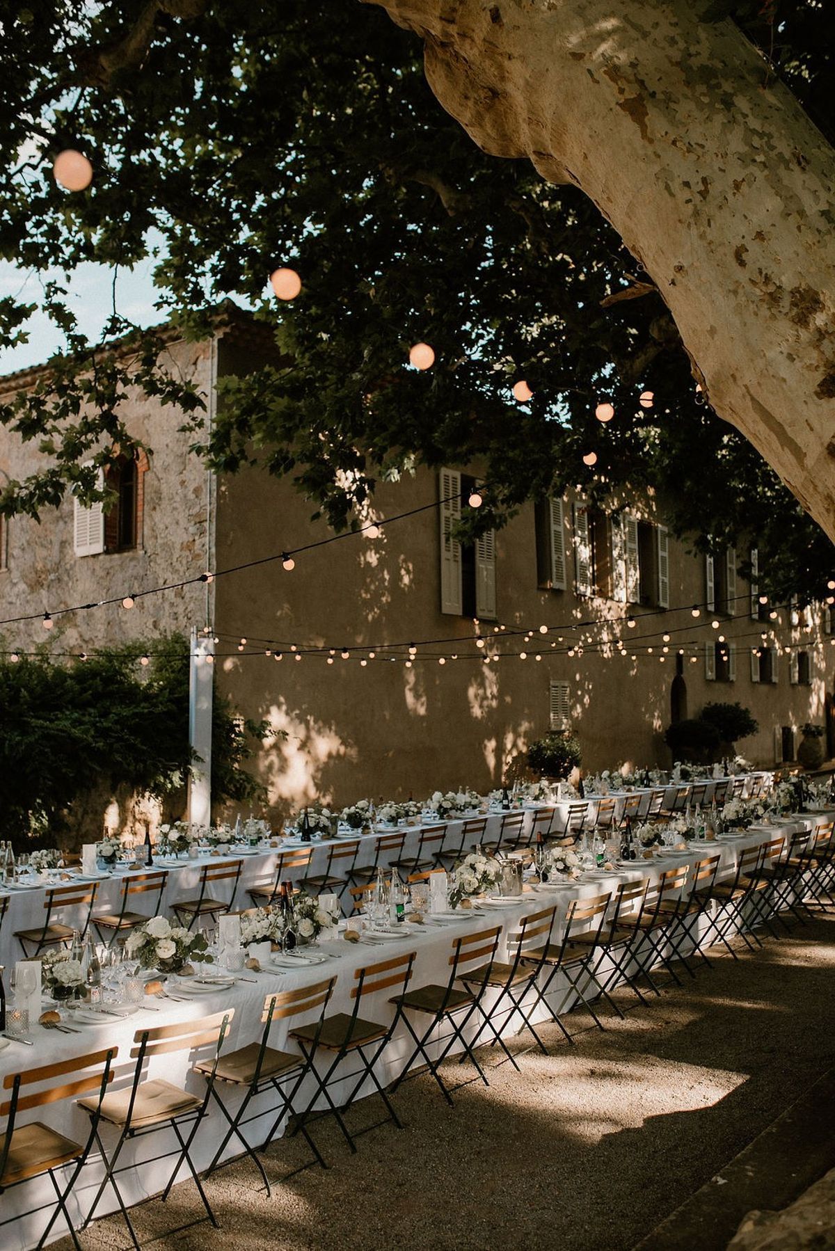Wood and metal bistro chairs set along banquet tables with festoon lights and chateau facade behind at Château Sainte-Roseline
