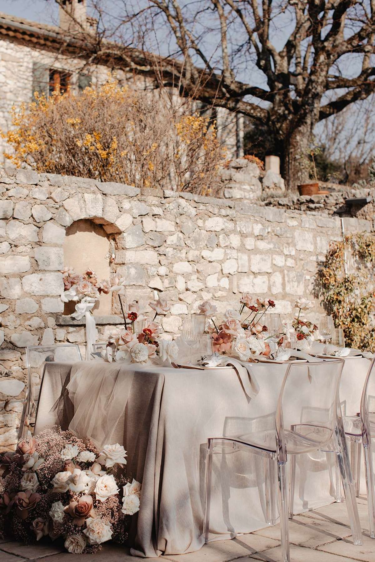 An outdoor reception table setup photographed in a courtyard against a rustic stone wall, with no people present. The long rectangular table is dressed in a greige linen tablecloth with a sheer tulle runner draping over the edge, and is surrounded by clear acrylic ghost chairs. The floral design features a cascading arrangement at the table's end and several centerpieces composed of ivory garden roses, blush and caramel-toned roses, deep burgundy ranunculus, blush gypsophila, and white magnolia-style blooms on dark stems. Place settings include ribbed glassware, gold flatware, and neutral-toned plates. The overall color palette is warm greige, blush, ivory, and terracotta, with a romantic yet modern aesthetic. This is a medium-wide portrait-orientation shot capturing the full length of the table.