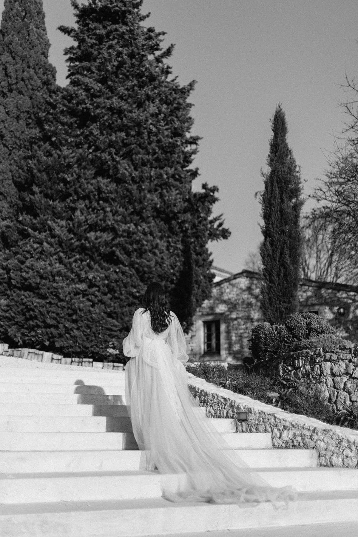 This black-and-white bridal portrait shows a bride photographed from behind as she walks up a wide stone staircase toward a stone building with tall windows. She wears a wedding dress with voluminous bishop-style sleeves and a long flowing tulle skirt that trails several steps behind her, with a large bow detail at the back waist. Her dark hair falls loosely over her shoulders. The outdoor setting features tall cypress trees, manicured boxwood shrubs, and a low stone retaining wall, suggesting a formal French or Mediterranean estate garden. The image has strong contrast between the bright white staircase and the deep dark tones of the trees, with soft mid-tones in the dress. This is a full-length portrait composition shot from a low angle behind the subject. Potential venue feature image.