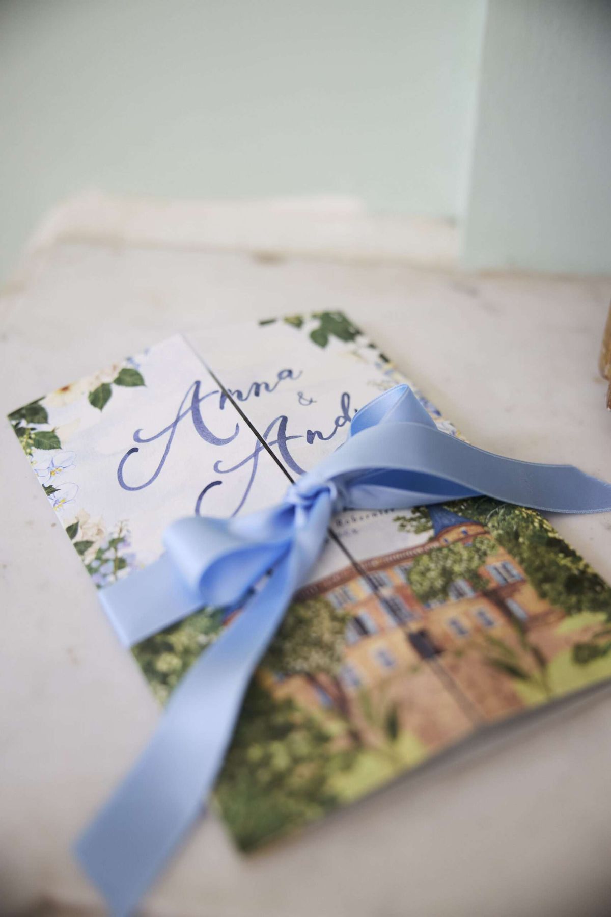 Close-up detail shot of a wedding stationery suite resting on a marble surface. The set includes two pieces tied together with a periwinkle blue satin ribbon bow: a ceremony booklet or order of service featuring hand-lettered calligraphy reading 'Anna & Andi' in navy blue ink with a white floral and green foliage border design, layered over a card printed with a watercolor illustration of a large ochre-toned manor or château surrounded by trees. The stationery palette combines navy blue script, soft blue ribbon, white blooms, and illustrated greenery, suggesting a classic yet painterly wedding aesthetic.