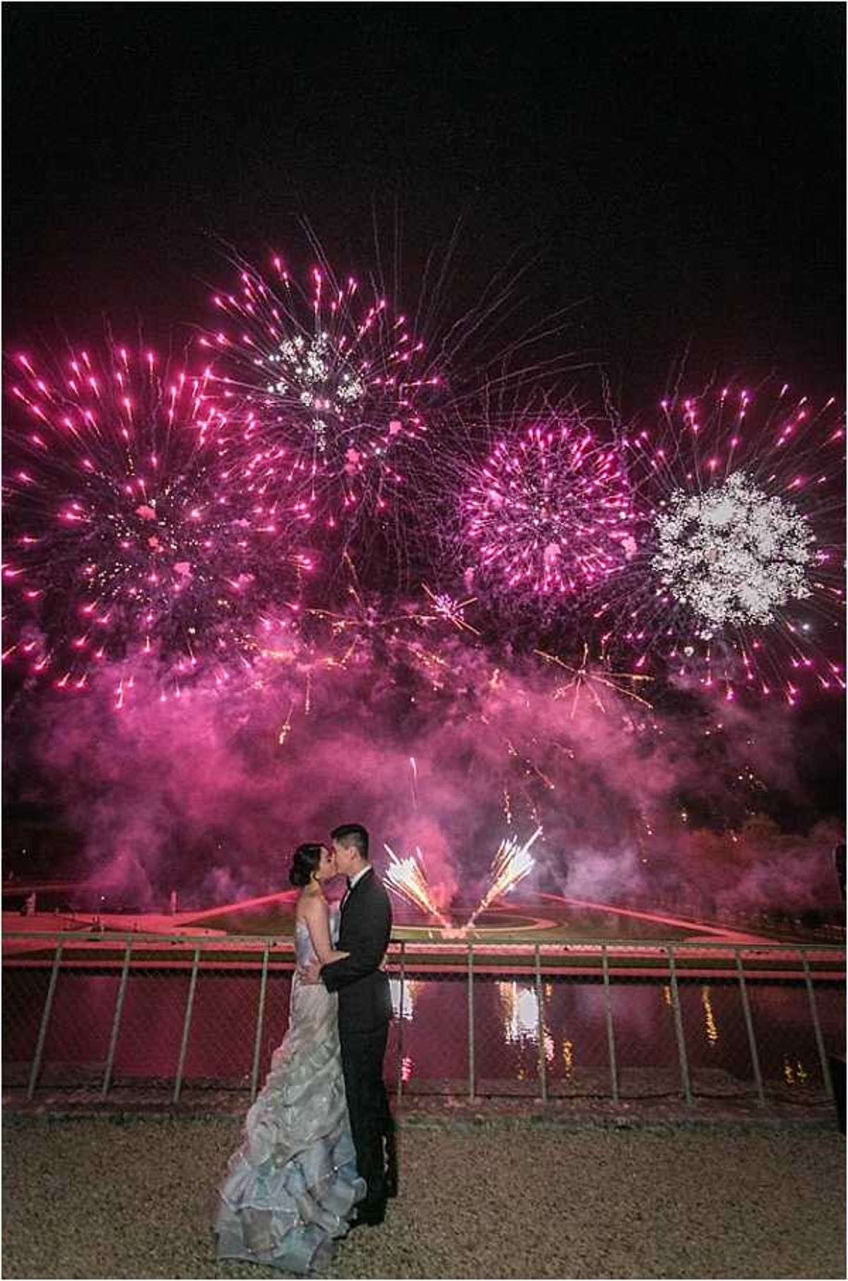 A couple shares a kiss outdoors at night in front of a large fireworks display bursting in vivid magenta, pink, and white tones against a dark sky. The bride wears a tiered, ruffled ball gown in pale icy blue-grey, while the groom is dressed in a dark suit with a white dress shirt. They are standing on a gravel path in front of a metal railing, with a reflective ornamental pond or canal visible behind them; ground-level firework fountains are also firing in the background, forming a heart-like arc. The wide-angle portrait composition places the couple in the lower third of the frame, with the fireworks dominating the upper two-thirds and their reflections visible in the water below.