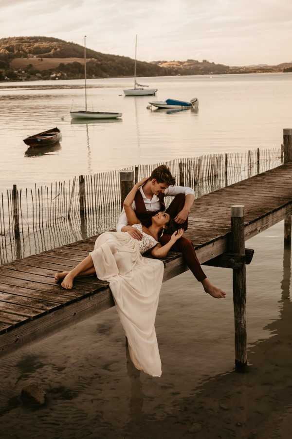 Barefoot couple reclines on weathered dock at dusk with moored sailboats in golden-amber light