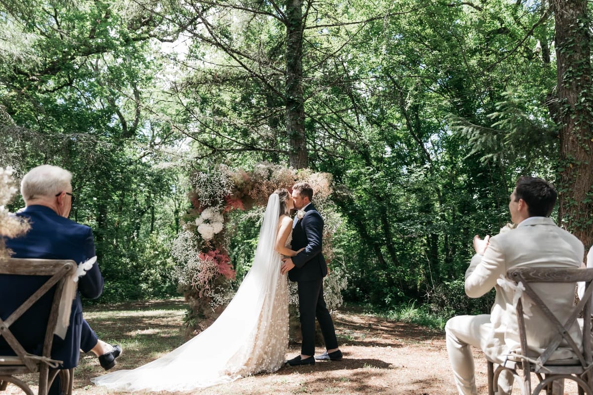 First kiss under circular floral arch in woodland clearing with cross-back chairs and guests
