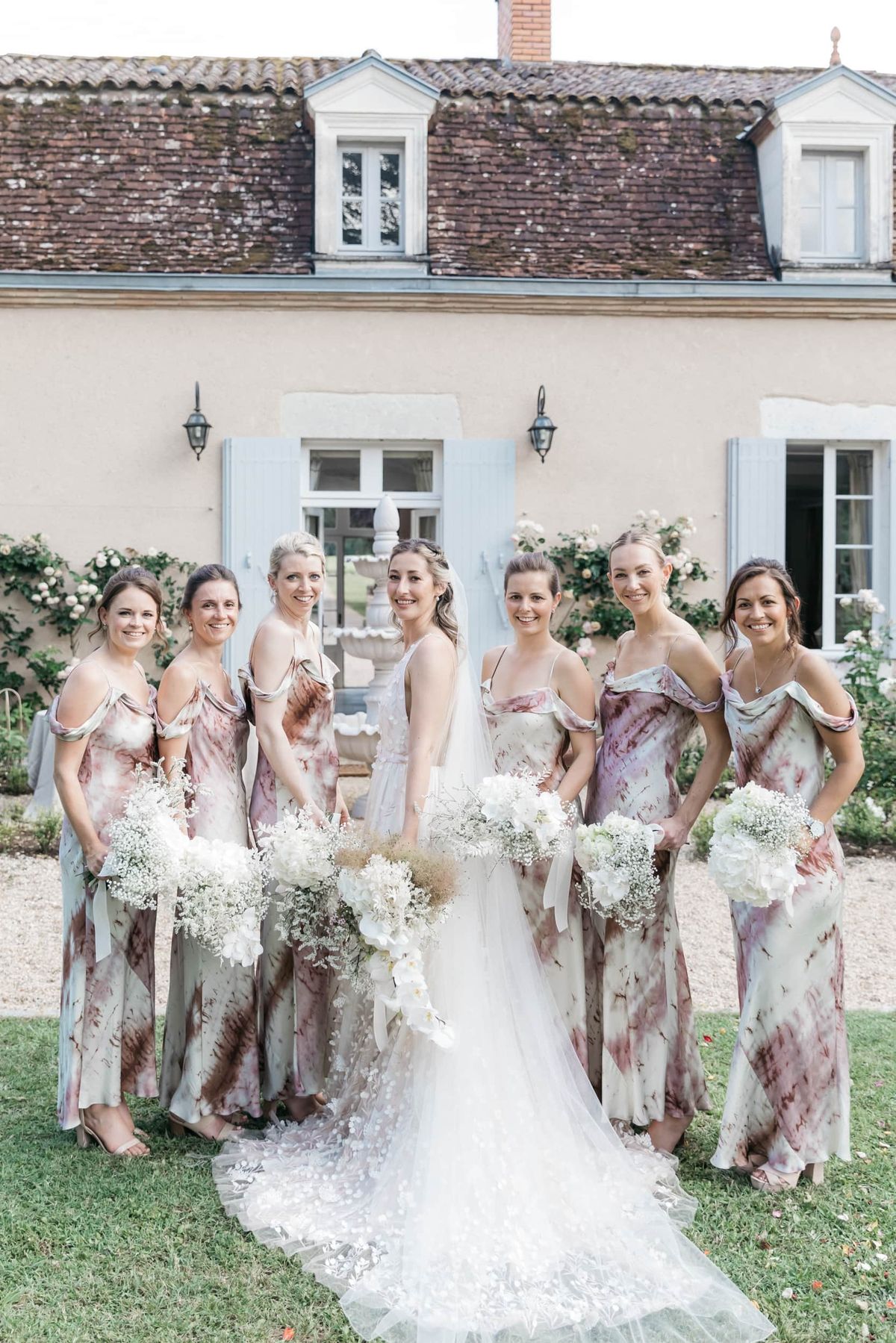 A bridal party portrait taken outdoors on the grounds of a French château or manor house, featuring the bride and six bridesmaids posing together on a lawn. The bride wears a white lace gown with a long cathedral-length embroidered tulle train and a veil, and carries a loose white bouquet featuring white orchids, dried grasses, and soft white blooms. The six bridesmaids wear floor-length tie-dye slip-style dresses in muted tones of dusty mauve, sage green, and taupe, with off-shoulder or cold-shoulder detailing, each carrying rounded bouquets of white hydrangeas and baby's breath. The venue building behind the group features pale yellow rendered walls, pale blue shutters, a terracotta tile roof with dormer windows, and climbing roses. The composition is a mid-range group portrait shot in natural daylight.