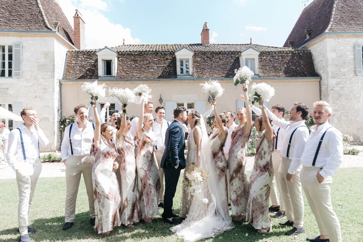 A bridal party group portrait taken outdoors on the lawn in front of a French château with limestone walls, terracotta roof tiles, and pale blue shutters. The couple kisses at the center — the groom in a navy suit and the bride in a white gown with a long veil, holding a loose bouquet of dried and white florals. Surrounding them are approximately five bridesmaids wearing matching floor-length tie-dye dresses in a cream and rust/burgundy marble pattern, raising bouquets of white baby's breath and white hydrangeas in the air. The groomsmen and male attendants are dressed in white shirts with beige or white trousers, several wearing navy or blue suspenders, and they are cheering and laughing. The overall styling is relaxed and modern with a boho-influenced color palette of white, cream, and rust tones. Wide shot with the château façade centered in the background. Potential venue feature image.