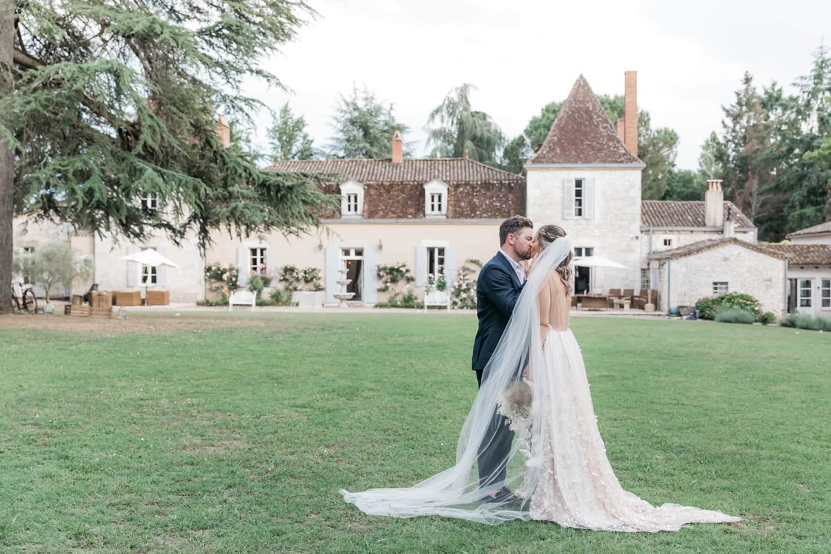 Bride in blush ballgown with cathedral veil kissing groom on lawn before multi-building chateau estate