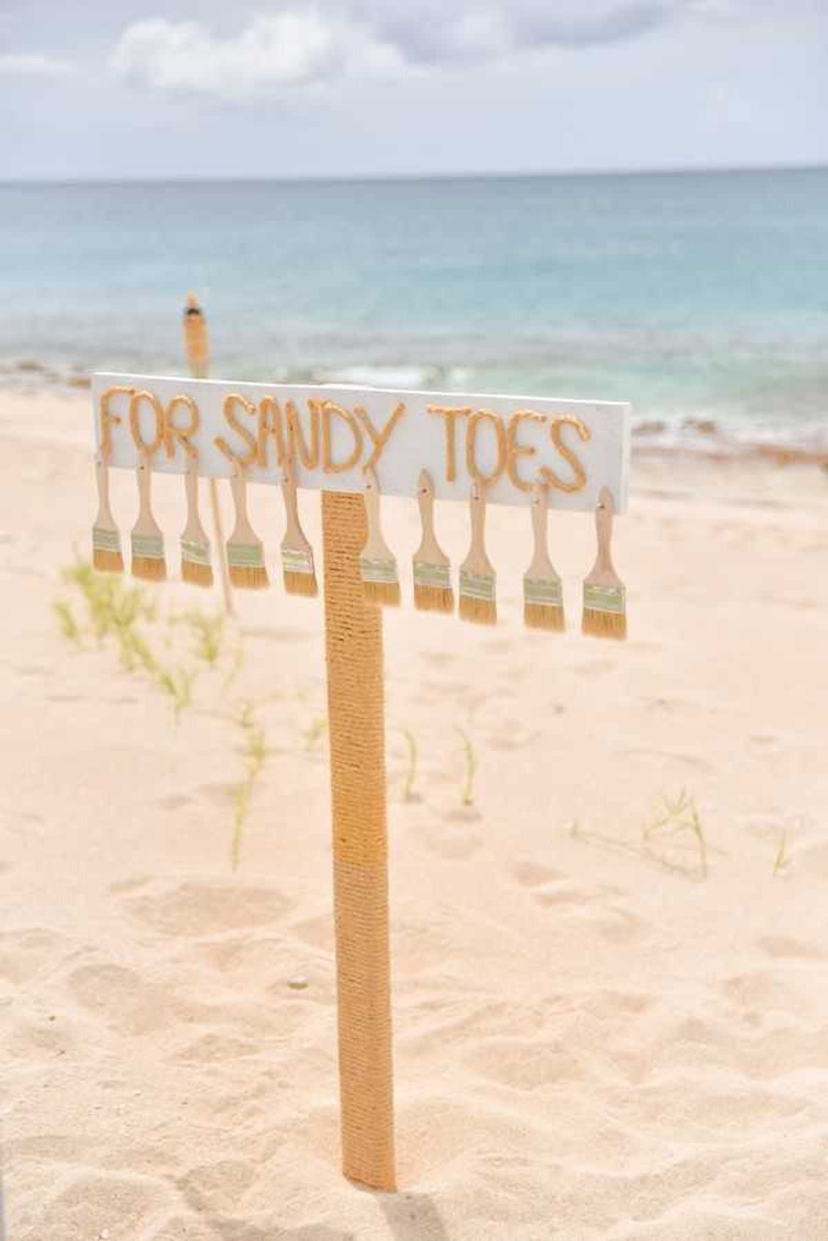 White wooden sign reading For Sandy Toes with brushes attached, planted in sand near turquoise water
