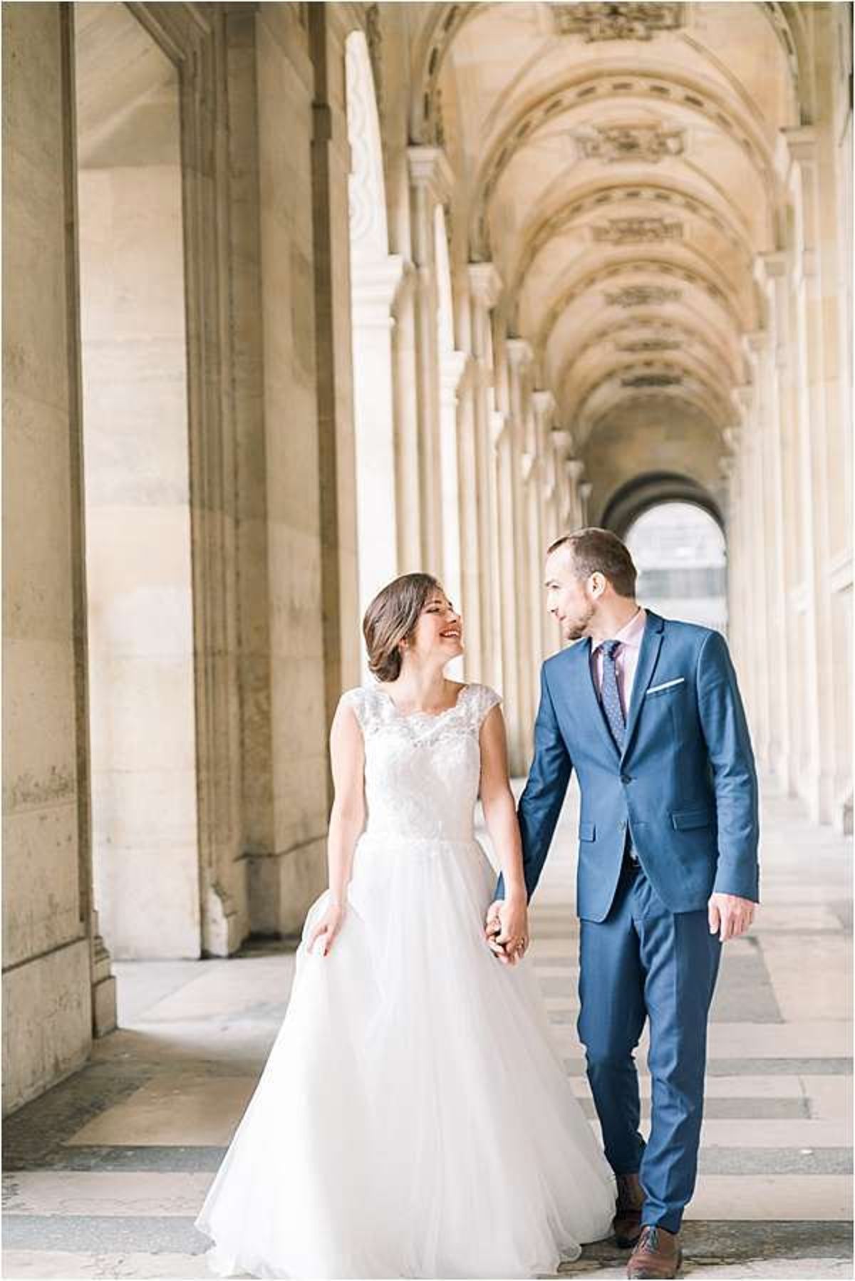 A couple walks hand-in-hand through a grand Parisian colonnade, laughing and looking at each other. The bride wears a white A-line gown with a lace appliqué bodice and a flowing tulle skirt, while the groom wears a fitted navy blue suit with a mauve patterned tie and a white pocket square. The setting is an ornate covered arcade with tall stone columns and vaulted decorated ceilings, consistent with the arcades of the Louvre or the Rue de Rivoli in Paris. The portrait is a medium full-length shot with the colonnade's repeating arches providing strong depth and perspective in the background.