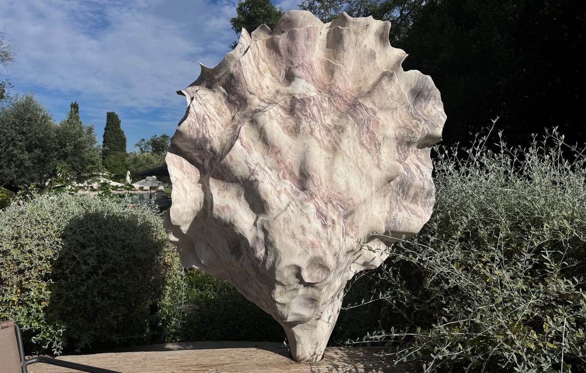 This image shows a large sculptural art piece positioned outdoors in a manicured garden setting. The sculpture is cream and pale pink with mauve veining, shaped like an oversized abstract organic form resembling a giant shell or leaf, and appears to be made from marble or a marble-effect material. It sits on a weathered wooden surface, with trimmed hedges, olive trees, and cypress trees visible in the background, along with what appears to be a pool terrace area with white umbrellas in the distance. No people, wedding party, or wedding-specific decor are present in this image — it is a garden sculpture detail shot with no clear wedding content.