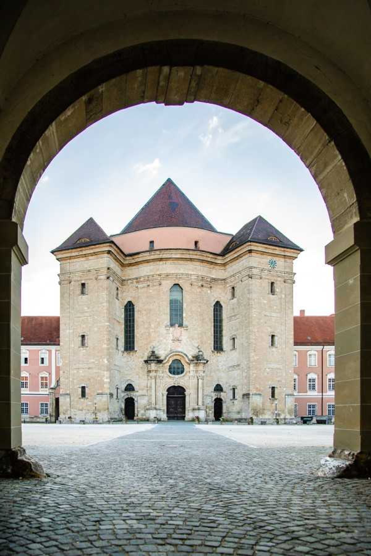 Baroque chapel facade framed through a stone archway with cobblestone courtyard and flanking pink buildings