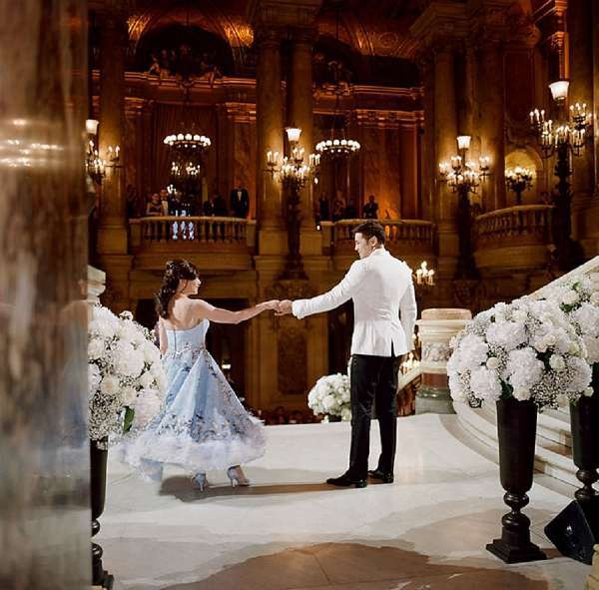 First dance at Palais Garnier staircase bride in blue tea-length gown groom in white jacket white hydrangea urns flanking