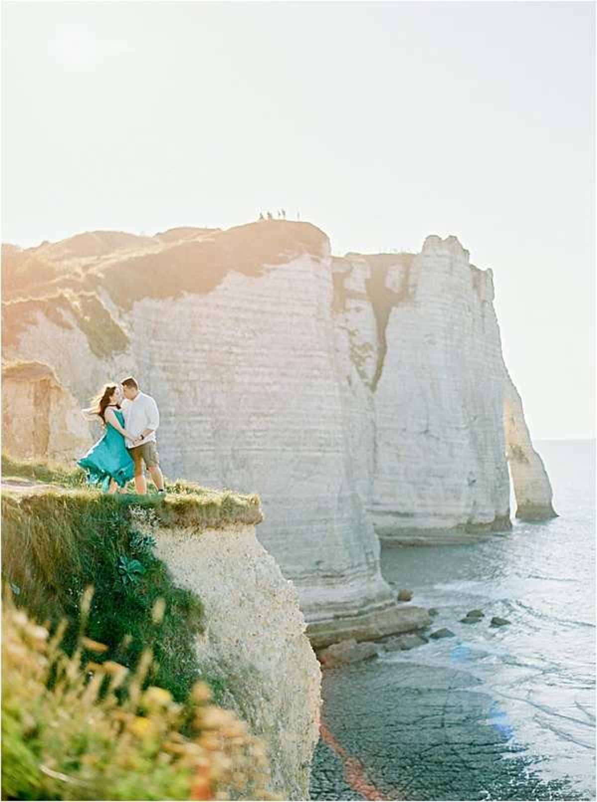 Couple on Etretat chalk cliffs woman in teal dress man in white shirt with dramatic white cliff arches and sea behind