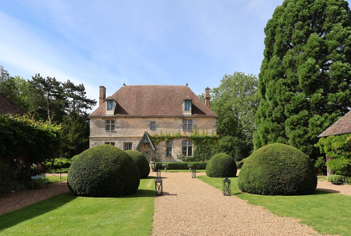 Stone manor with timber-framed porch, mansard roof, topiary box hedges, and lantern-lined gravel drive