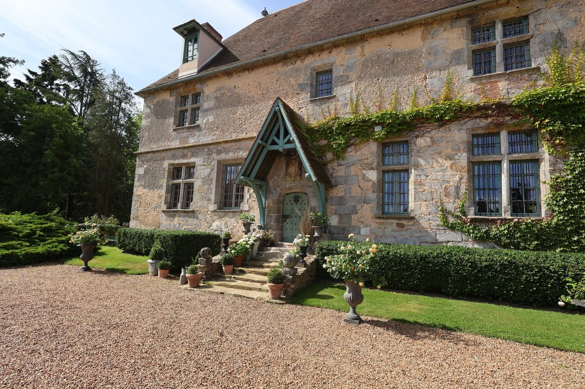 Exterior wide shot of a historic French stone manor house with no people visible. The building features a distinctive pointed entrance porch with sage green painted timber framing, a matching sage green door, and multi-pane windows with the same green trim throughout. Climbing vines spread across the upper right facade. The entrance steps are flanked by terracotta pots, dark grey stone urns with white hydrangeas and peach-toned roses, and clipped boxwood hedging. A gravel forecourt leads up to the entrance. The architecture is classic French country manor style, two stories with a steep tiled roof and a small dormer window. Potential venue feature image.