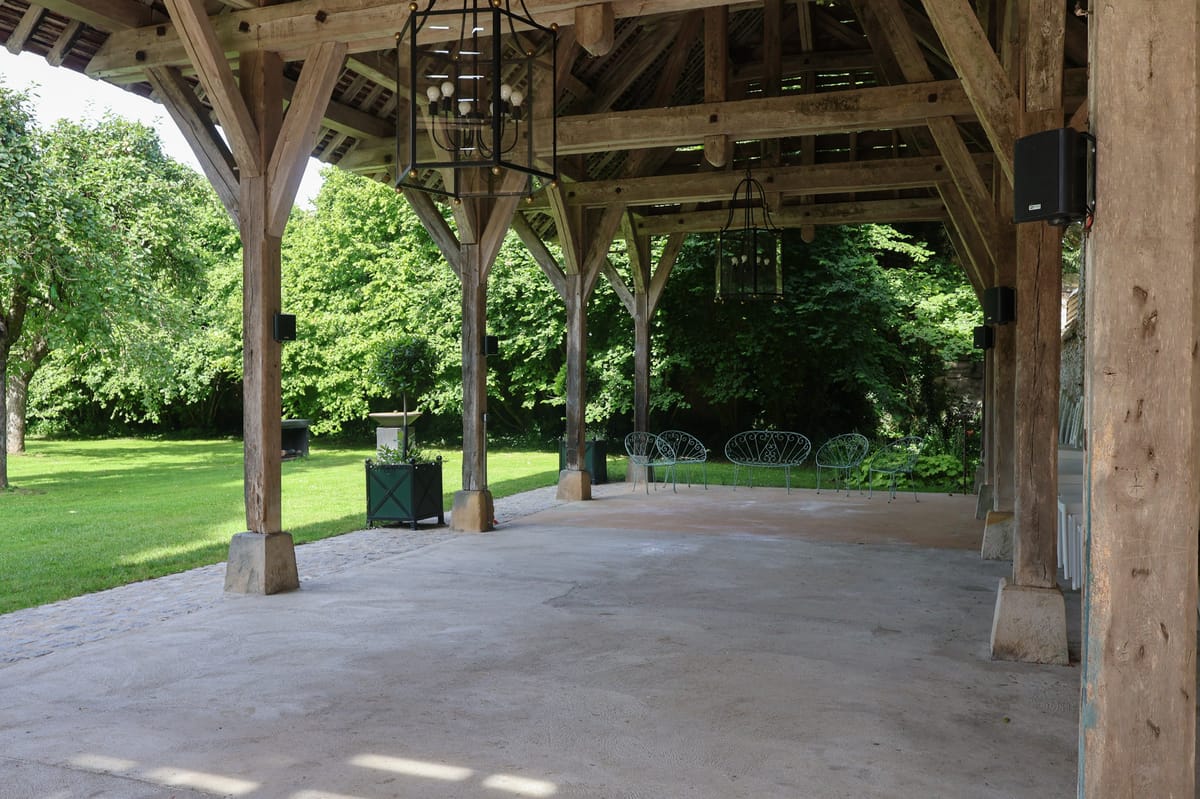 Open-sided timber-frame pavilion with rustic wooden beams, wrought-iron chandeliers, and wrought-iron garden furniture