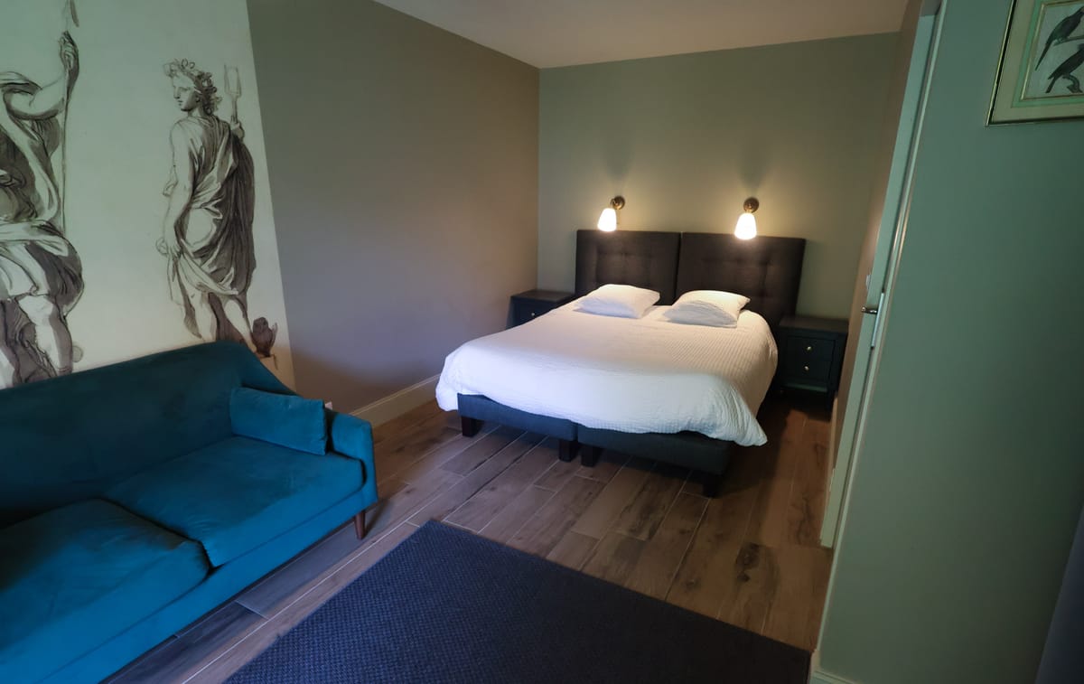 This image shows a guest bedroom, likely part of a wedding venue's accommodation offering. The room features a double bed with a dark charcoal upholstered headboard, white bedding, and two matching dark navy bedside tables with warm-toned wall-mounted sconce lights. A teal velvet sofa sits in the foreground beside a large feature wall panel depicting a classical figure rendered in a grey illustrative style. The walls are painted in a muted sage-grey tone, the floor is light oak-effect wood, and a navy blue area rug is partially visible. A framed botanical bird print is visible on the right wall near an open door. The room's decor palette combines teal, navy, and warm grey in a classic-contemporary style. Wide-angle interior shot. Potential venue feature image.