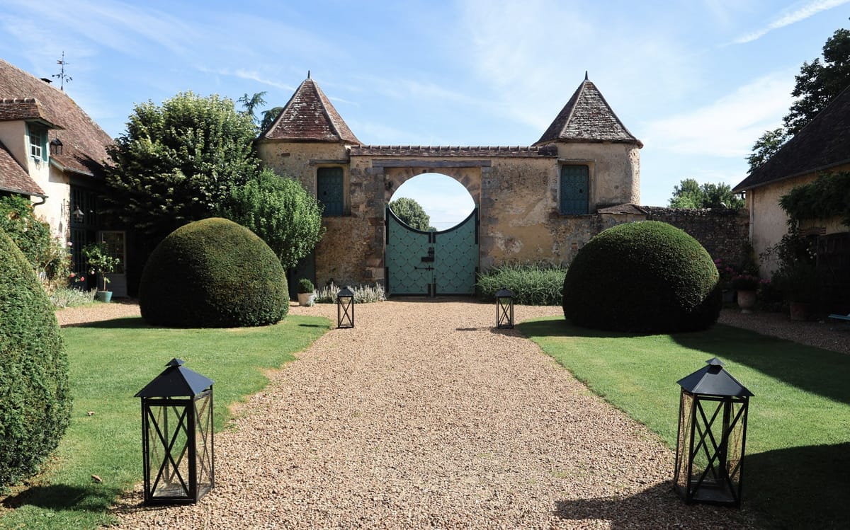 Symmetrical gravel path to stone gatehouse with twin conical turrets and teal doors flanked by topiary