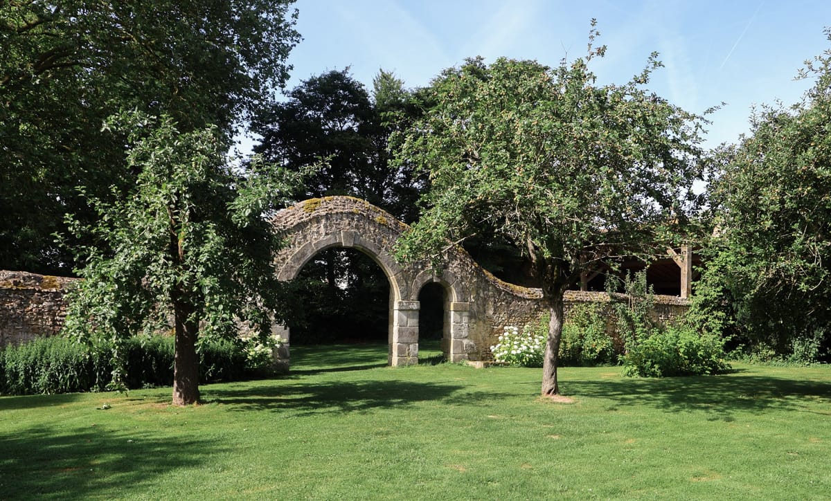 Romanesque stone archway in moss-covered wall with white hydrangeas and wooden pergola beyond