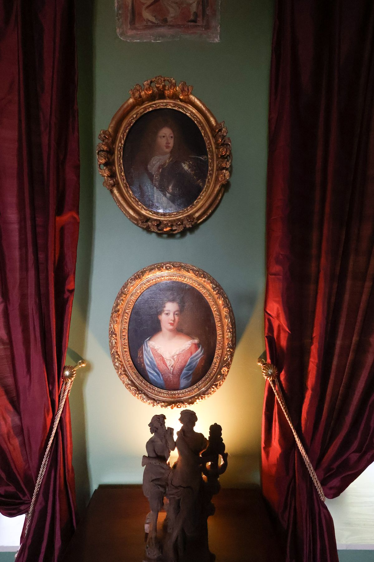 An interior detail shot of a château or historic manor venue, showing a pale sage-green wall flanked by floor-length deep burgundy silk curtains tied back with twisted cord and gold tieback hooks. Two oval oil paintings in ornate gilded frames hang vertically on the wall — the upper portrait depicts a man in dark armor, the lower depicts a woman in a red and blue décolleté gown with auburn hair. At the base of the wall, two stone or plaster figurative sculptures are illuminated from below by a warm accent light, casting a golden glow against the wall. No people or wedding party are present. Potential venue feature image.